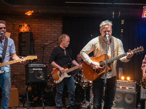 Full band on stage in a brick-wall bar with EXIT sign, warm lamp lighting
